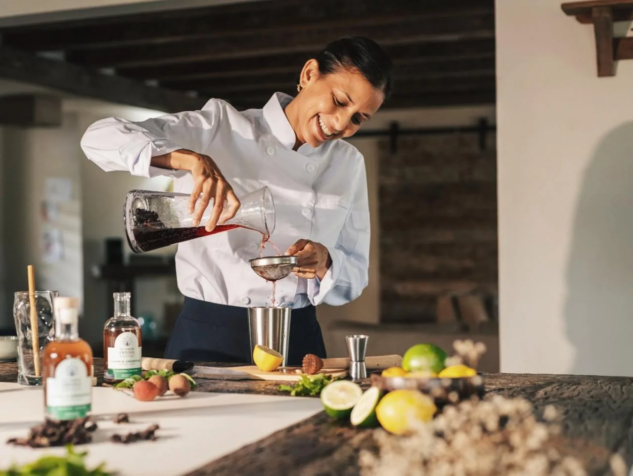 Tessa pouring a freshly made Le Verger Créole cocktail, syrup bottles and tropical fruit on the counter